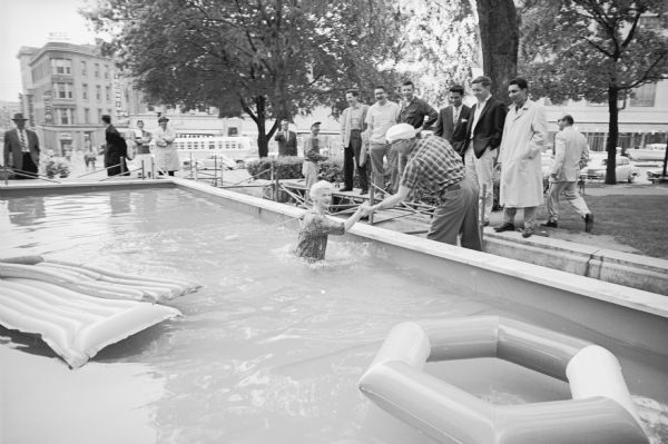 Model Marlyn Sachjen being helped out of the temporary pool which had been constructed on Capitol Park at the head of State Street. She had been modeling summer sportswear while paddling around on a rubber raft when she fell into the water after the raft overturned. The "Outdoor Living Day" event was sponsored by the Madison Chamber of Commerce.    