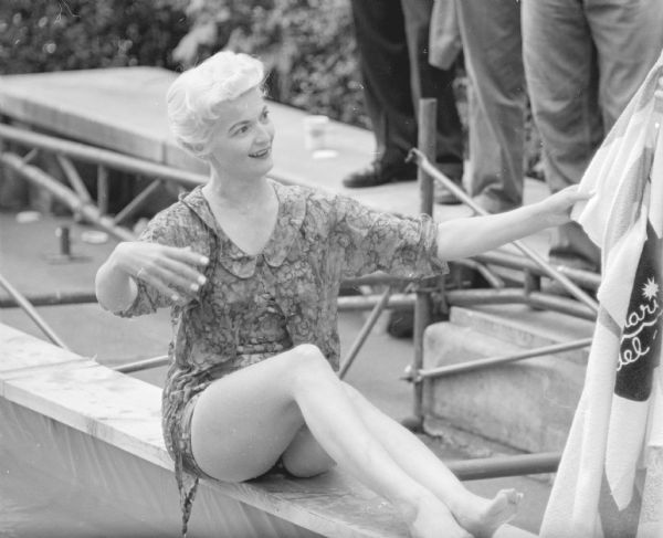 Model Marlyn Sachtjen sitting on the edge of a temporary pool that had been installed on Capitol Park. She is wet after having fallen off a floating raft while modeling summer sportswear. She is reaching for a towel that was part of the display. The "Outdoor Living Day" event was sponsored by the Madison Chamber of Commerce.  