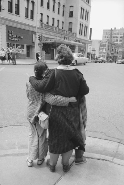An unidentified woman hugging two children while standing on the Capitol Square across the street from the Park Hotel. At the corner of the Park Hotel on the left is The Prescription Pharmacy.