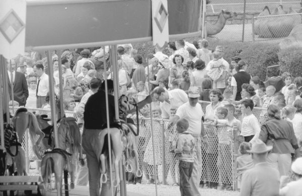 Children and parents waiting their turn to ride the new Vilas Park merry-go-round during the grand-opening event for the ride on June 21, 1958. In the background are camels behind a chain-link fence.