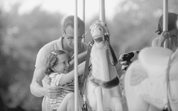 A young girl taking her first ride, with assistance from her father, on a horse on the new merry-go-round in Vilas Park. The grand opening for the merry-go-round occurred on June 21, 1958.