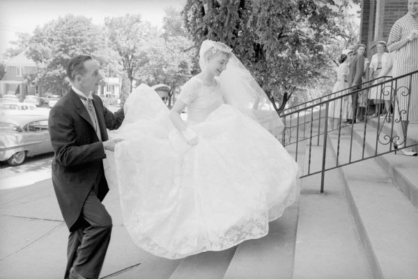 As the bride, Susan Renk, is walking up the steps of the Sacred Hearts Catholic Church in Sun Prairie, her father, Wilbur Renk, is assisting by holding up her skirt and veil.