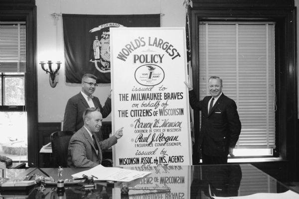 Three men next to a large poster with the words: "World's Largest Insurance Policy issued to Milwaukee Braves on behalf of Citizens of Wisconsin. Vernon Thomson, Governor. Paul J. Rogan, Insurance Commissioner. Issued by Wisconsin Association of Insurance Agents."