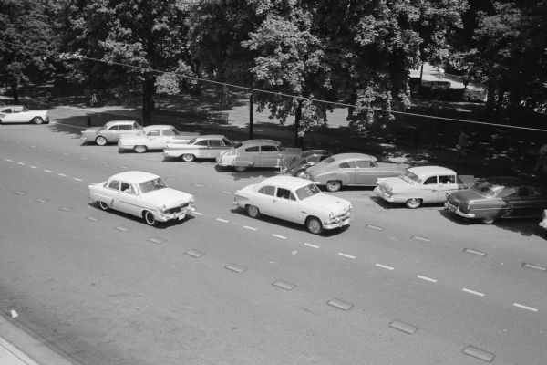Elevated view showing newly applied, experimental, white plastic traffic strips installed on the streets around the Capitol Square. The automobiles are traveling within the traffic lanes, and other automobiles are diagonally parked on the Capitol side of the street.