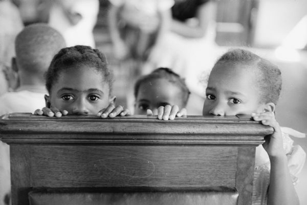 Three African-American pre-school girls peer out from behind a church pew at summer bible school.