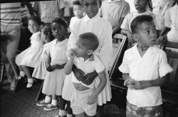 A group of children attending Bible school. In the foreground a 3 year old boy is sucking his thumb as he is leaning into the embrace of an older sister behind him.