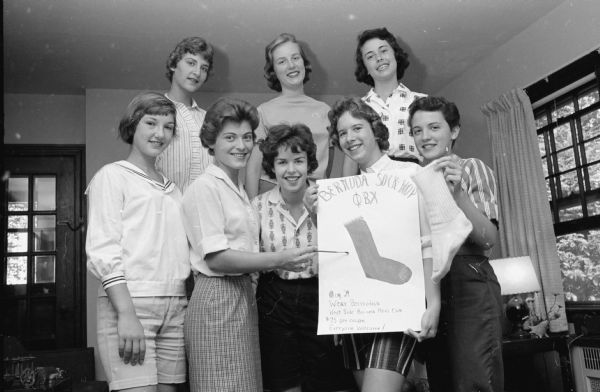 Sorority members planning the Phi Beta Chi "Bermuda Sock Hop". Front row, left to right, are: Marian Amlie, Pat Patterson, Kathleen Gorman Cathy Cline, and Barbara Berger. Back row, left to right, are: Elizabeth Stiehm, Jane Taplick, and Kathy Druckenbrod. They are posing around a homemade sign with a drawing of a sock in the center that reads: "Bermuda Sock Hop &#934;&#914;&#935; / Aug 29 / Wear Bermudas / West Side Business Men's Club $.75 per couple / Everyone Welcome!". A woman on the right is holding up a sock.