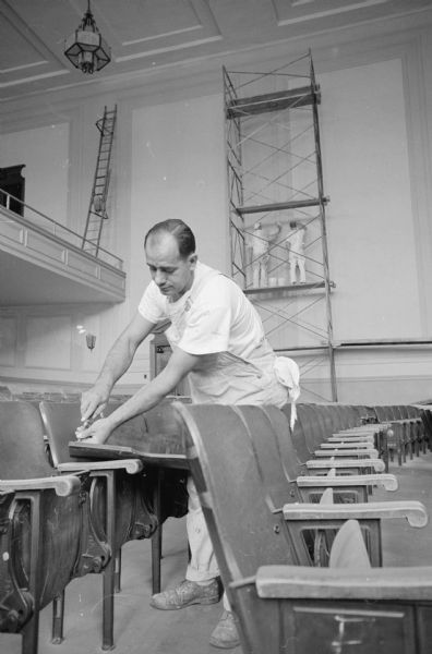 Ernest LaBella, Capital Decorating Co., is repairing a seat in the West High School auditorium as part of an effort to prepare city school buildings for the beginning of the school year. In the background are two people working on a scaffolding set up against the wall. A ladder is set up on a balcony on the left.