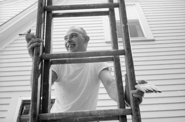 Norbert Anderson friends who pitched in to help paint his home at 1522 Jefferson Street. View looking up at Roland Wiessinger, who holding a cigarette in one hand and a paintbrush in the other while moving a ladder. The side of a house is in the background.