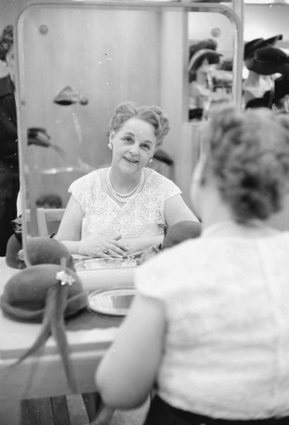 The original caption states: "Looking at her reflection in a mirror is Irene Hubbard, president of the auxiliary to Local 1404, United Steel Workers of America (AFL-CIO). Her husband Paul Hubbard is a machinist at Gisholt Machine Co. She is a saleslady in millinery at W. J. Rendall's store."