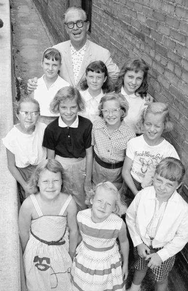 Group portrait of ten girls, who called themselves the "Mifflin Moppets" posing with <i>Wisconsin State Journal</i> sportswriter Joseph "Roundy" Coughlin in the backyard at 1307 E. Mifflin Street where they held an event to raise money for Roundy's Fun Fund, a charity which benefitted children with disabilities.