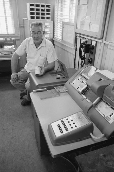 R. E. Gilbert posing behind a desk-sized IBM machine, known as a integrated-data processor. Gilbert holds in his hand a punched paper roll containing up-to-the-minute information about the freight cars in the Madison North Western railroad yard. The information is relayed to the Chicago office where it can be shared almost immediately.