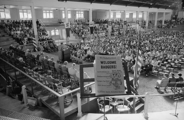 Wide angle view of 2,700 freshmen students in the Stock Pavilion being officially welcomed to the university. In the foreground is a large sign on an easel that reads: "Welcome Badgers!" with an image of Bucky Badger.