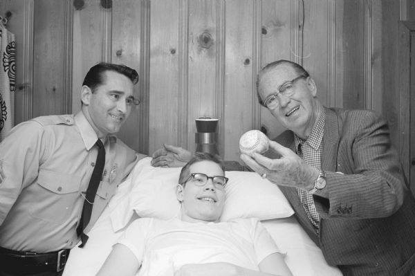 Mike Falconer (17), 626 Eugenia Avenue, an avid Milwaukee Braves fan, receives an autographed baseball from the team. Mike, son of Mr. and Mrs. H. B. Falconer, suffered a spine injury in a fall at Medford, WI in April 1957 which paralyzed him from the waist down. A West High senior, Mike takes instruction daily via a telephonic setup at his bedside. Shown (L-R) are: Madison Police Sgt. Robert L. Ferris, Mike's neighbor; Falconer; and Joseph (Roundy) Coughlin, who delivered the ball on Mike's birthday.