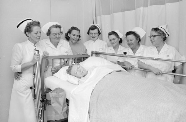 Mrs. Anna McFarlane is surrounded by nurses in her bed on the newly remodeled 4th floor of the Lake View Sanatorium. Mrs. McFarlane is surrounded by attendants, (L-R): Mrs. Janette Ditsch, Loella Karow, Mrs. Opal Degler, Doreen Steensrud, Mrs. Ivy Gunsonlus, Mrs. Mildred Zimmermann, and Mrs. Myrta Kerrigan.  