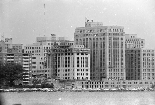 View across Lake Monona towards the third and final wing to the Wilson Street state office building. Automobiles are parked along the shoreline.