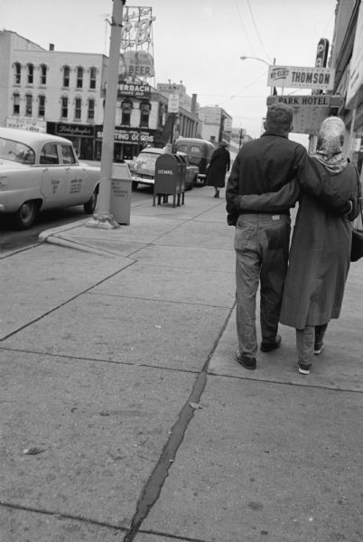 A couple walking on the sidewalk alongside North Carroll Street towards the Park Hotel. Storefronts across the intersection on the left along Main Street include Forbes-Meagher, and Wisconsin Felton Sporting Goods.
