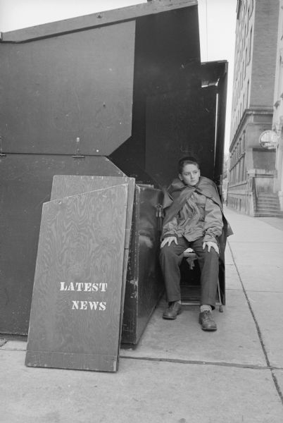 An attendant at a newsstand in downtown Madison. A sign reads: "Latest News." A sign on a building along the sidewalk on the right reads: "Hob Nob Bar".