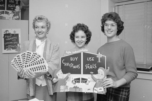 One in a series of photographs depicting volunteers preparing a mailing of Christmas Seals as part of a campaign to fight tuberculosis. Virginia Bowen, left, is displaying sheets of the 1958 seals. Cam Mazer, center, and Helen Wagner are holding a poster urging citizens of Madison to buy and use the seals, which cost one cent apiece. 