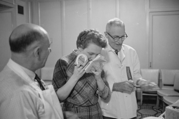 <i>Wisconsin State Journal</i> reporter, Marcia Crowley, at a cheese display at the 67th annual meeting of the Wisconsin Cheese Makers Association. She is sampling camembert cheese. The two men with her are likely Horace Mulloy, field manager of the association and Ernest Jung, superintendent of the exhibits. 
