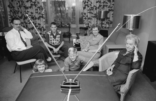 Three generation family watching television, shot from behind the television through the 'rabbit ears' antenna. At the far right is 'Granny' Mary B. Cowden, holding in her ear 'a special gadget which helps her to hear the sound better'. Shown left to right, are: Ralph Metz; Phyllis, 9; Allen, 15; Scott, 12; Mary C. Metz and Cowden.  