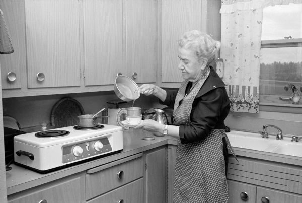Mary B. Cowdin is standing by a two-burner stove, and pouring hot water into a 'one cup' coffee maker. She resides in her own apartment within her daughter's family's house.