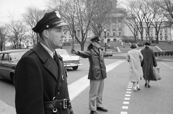 Cadet Policeman Daniel Writt is shown receiving training and directing traffic at the State Street corner of Capitol Square, while in the foreground, Donald Kjin, a veteran traffic officer, supervises. The class of 20 police cadets studied and trained for 16 weeks.  