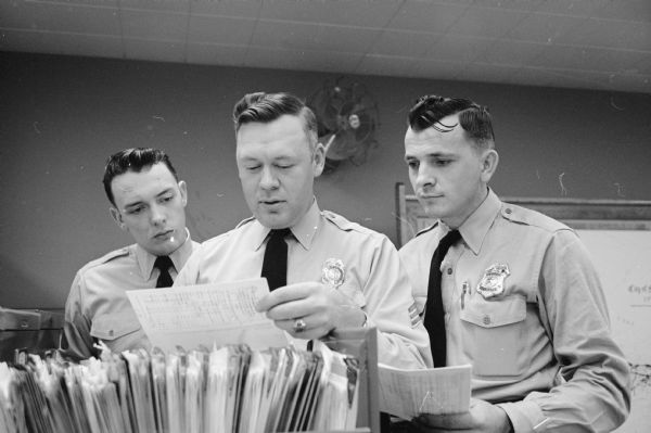 A policeman and two police cadets examine a large file of folders and papers containing warrants to violators who ignore parking and traffic arrest tickets. The cadets were Gerold Thorstenson (left) and Paul Zajicek (right). The policeman was Sgt. Hiram (Buck) Wilson (center).  