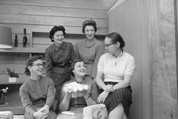 Group portrait of five members of the Dane County Dental Auxiliary, with one of them displaying a tiny lace Christmas tree 'skirt' which will decorate the group's annual scholarship dinner dance. Proceeds from the party will go to a scholarship fund for dental students. Seated are Lois Cardinal, Virginia Nania and Francis Linn. Standing are Harriet Kelly and Phyllis Jacobs. The five women are wives of dentists.