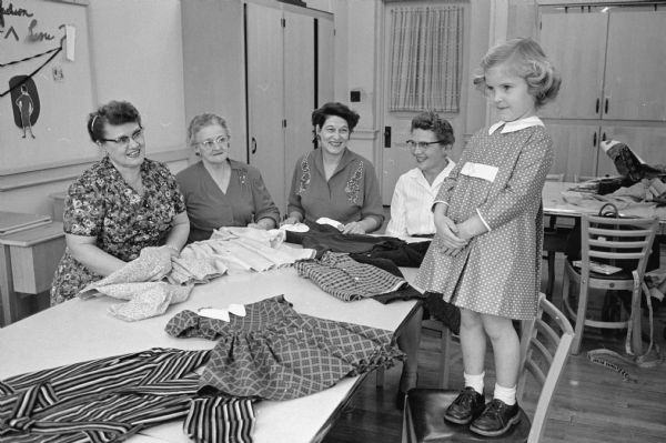 Group of grandmothers from a clothing class at Madison Vocational and Adult School that make clothing for their grandchildren. Genevieve Sorenson (left) admires her granddaughter who is modelling a dress made for her. Other women in the group, left to right, are: Marie Ludden, Dorothea Frye, and June Sime.