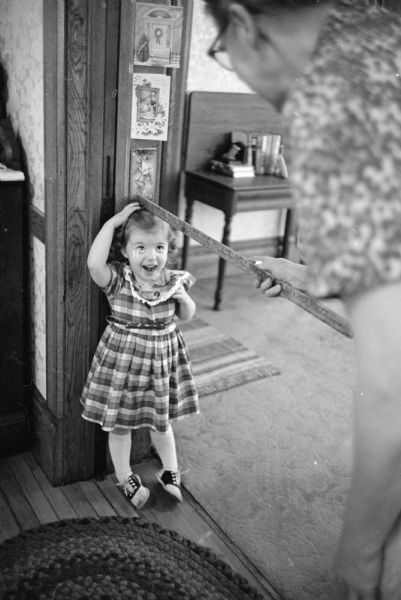 Observing a family tradition that the height of all children are measured each Christmas season on the sliding oak door between the parlor and dining room, Ann Denise, 3, is being measured by her grandmother. One in a series of images depicting Christmas activities at the 120-acre farm of Mr. and Mrs. Dennis Bayles, Rt 2, Lodi.