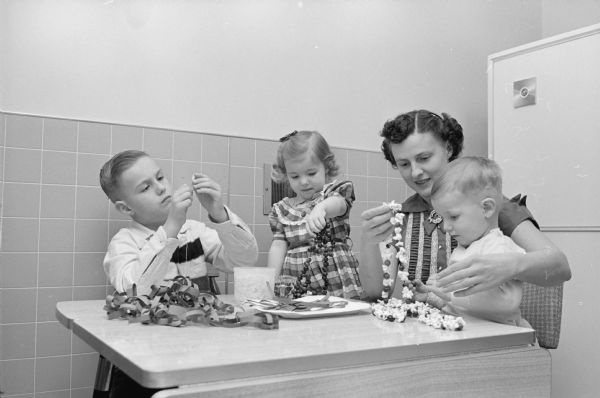 Jean (Bayles) Kirking instructs three of her children, Duane, Ann, and Bruce in the technique of stringing popcorn and cranberries and paper chains to decorate their Christmas tree.