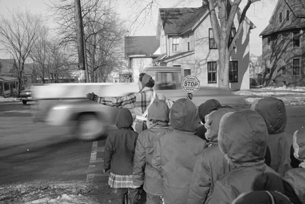 Crossing guard Meredeth Niemczyk, standing at the corner of Jenifer and Brearly streets, facing north, spreads her arms and displays a hand held stop sign to hold back nine children all with their hoods up, as a blurred pick-up truck races by.