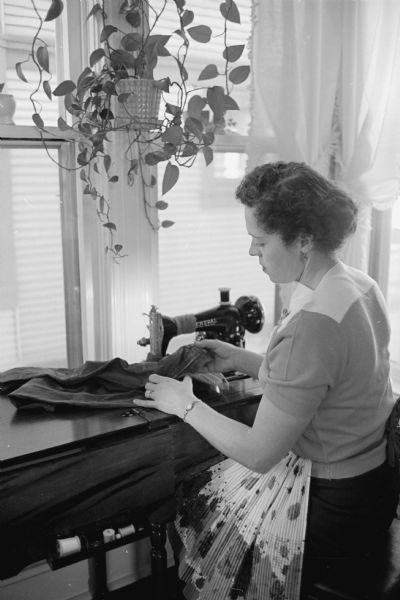 Crossing guard Meredeth Niemczyk is shown at home on break seated at a sewing machine by windows. The caption reads: "Despite a daytime routine that is interrupted three times by her crossing guard duties, Mrs. Niemczyk manages to find time to do the mending for her husband and two young sons."
