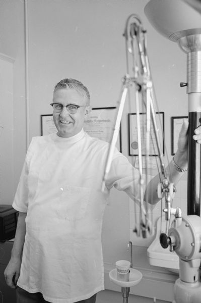Dentist Oliver Donkle is shown wearing a white work tunic and standing in his office by a folded dental drill with pulleys.