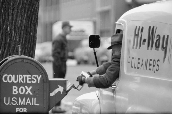 A man using two hands to deposit letters in a roadside mailbox from a truck on the corner of Monona Avenue and Doty Street. The photograph was taken with a long-lens camera.