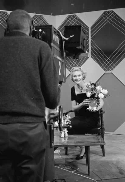 Beverly Stark, a Madison television personality with two children, is posing with a floral arrangement on a set in front of a television camera. A man is standing behind the WMTV camera in the foreground on the left.