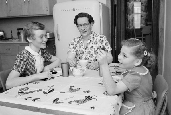 Private duty nurse, Mrs. Clayton R. Felland, sitting at a table at home with her two daughters, Chere and Candy.