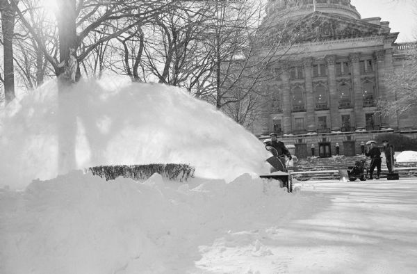 "What All Hope Snow Will Do: Blow" is the caption. As much snow on the ground as there was at anytime during the past two winters. A man is pushing the snow blower which is throwing snow high into the air. Two other men, one with a snow blower and one with a shovel, are standing at the base of the steps to the Wisconsin State Capitol building in the background.