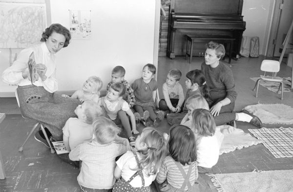 Young children paying attention to their teacher, Beverly McCumber, who is reading a picture book at a cooperative nursery school at Bashford Methodist Church. The nursery school is an experiment of the University of Wisconsin Dames club. Assisting is Marjorie Christianson, a member of the Dames club.