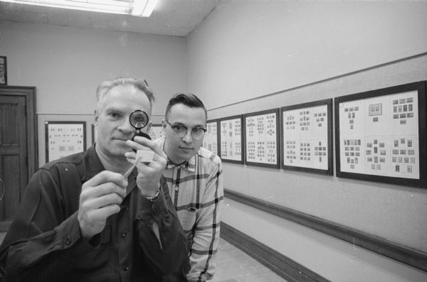 Members of the Madison Stamp Club shown examining a stamp. (Ned Lawrence on left, Fred Huebner on right). Displays of stamps on the wall behind them are part of the Stamp Club Annual Exhibit at the Madison Public Library.