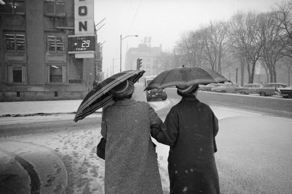 Two women, seen from behind, are using umbrellas while walking on a snowy sidewalk on the first block of East Main Street on the Capitol Square in Madison.