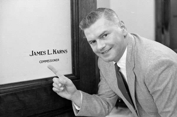 A man is pointing at his name and job title which were recently painted onto the frosted glass of his office door. The man was James L. Karns on his first day of his appointment as state motor vehicle commissioner.
