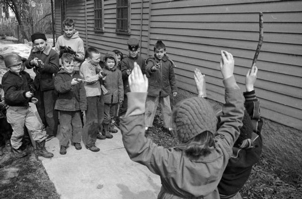 Two girls with their hands up being 'pretend' robbed by ten little boys with cap-guns. They are between two closely spaced homes.
