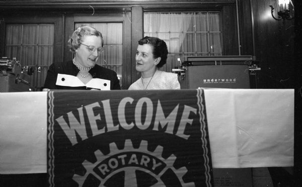 Two registration clerks confer behind a welcoming banner for the district Rotarian conference at the Hotel Loraine. They are Gertrude Fenner and Peggy Parriott. 