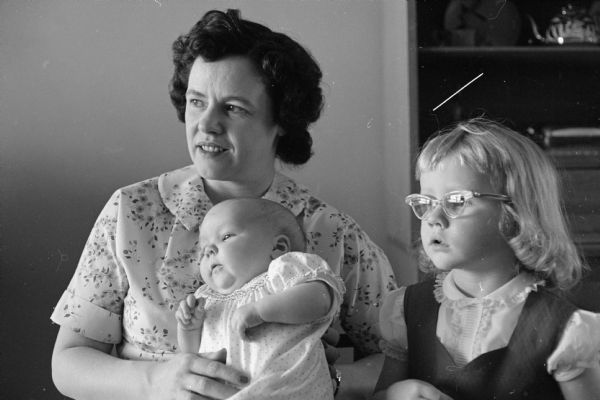 An unknown woman is holding a baby, and an unknown girl wearing glasses is looking on. Possibly Ed Stein relatives. Possibly Mrs. Skewis.
