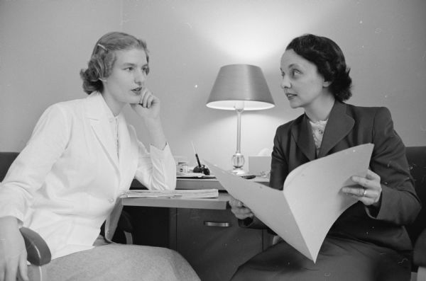 Two young women doctors who are completing their residencies in psychiatry at the University Hospitals, are shown discussing a case.  Seated on the left is Dr. Sonja Monson, and on the right is Dr. Frances E. Ehrlich.