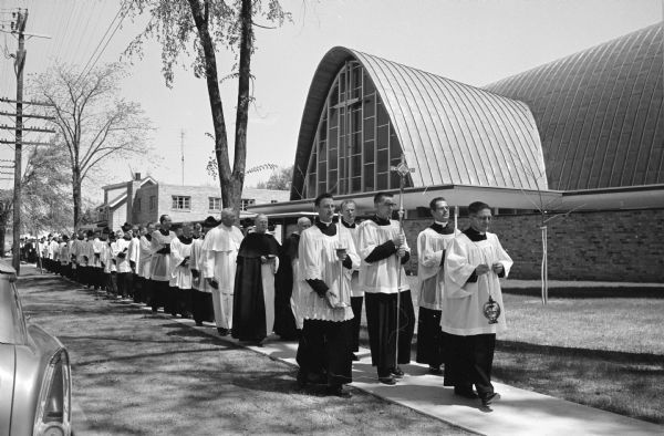 A procession of clergymen walks in front of the new Middleton St. Bernard Catholic Church on Parmenter Street in Middleton. The modernist building features an arched roof with a large cross and stained glass window.