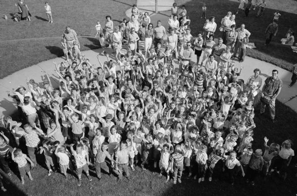 Overhead view of the children of Dudegon school on Monroe Street gathered on the front lawn for the annual school picnic. Men and women are standing on the outside of the circle of children, many of them waving at the camera.