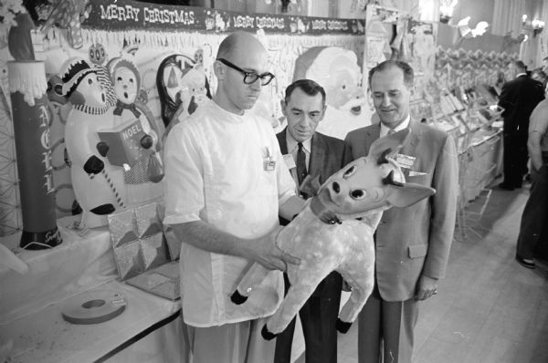 A man in a pharmacist smock is examining a large toy as two other men are looking on. They are at the Hotel Loraine at a merchandise display for all merchants who handle Rexall Drug Co. products. Behind them is a Christmas display. The men are (L-R) John Jacobson of the Rennebohm Drug Co., store #4; Harold Blum, operator of Blum's Rexall store, Monroe, Wis.; and Charles Rhoten, district Rexall manager, from Chicago.